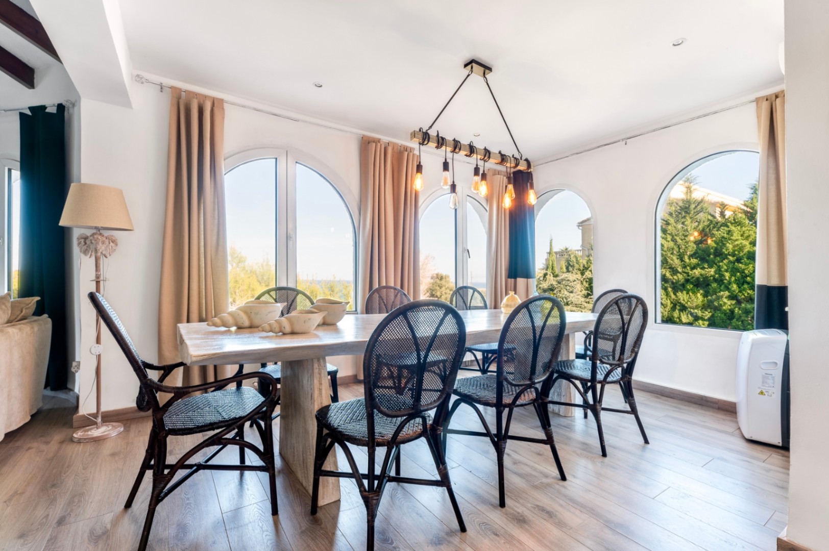 Comedor luminoso con mesa de madera rústica, sillas de mimbre negro y amplios ventanales en arco que ofrecen vistas al entorno natural de Jávea.