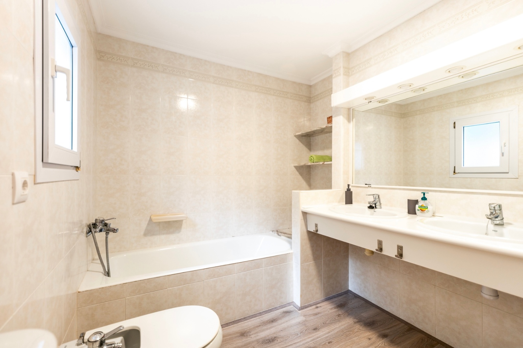 Cuarto de baño con bañera, bidé y tocador de doble seno. Incluye azulejos en tonos crema, espejo de gran formato con iluminación integrada y suelo de efecto madera.