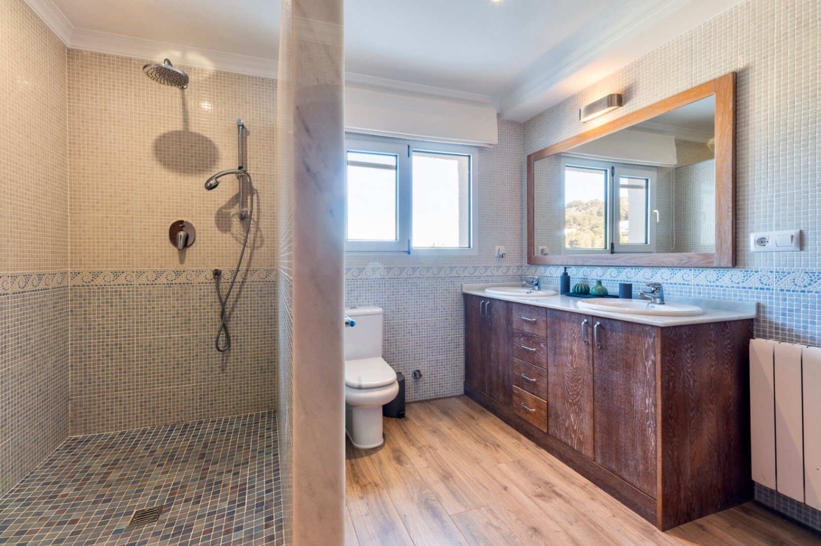 Cuarto de baño con ducha de mosaico, mueble de doble lavabo en madera oscura, espejo de marco rústico y suelos de efecto madera con luz natural.
