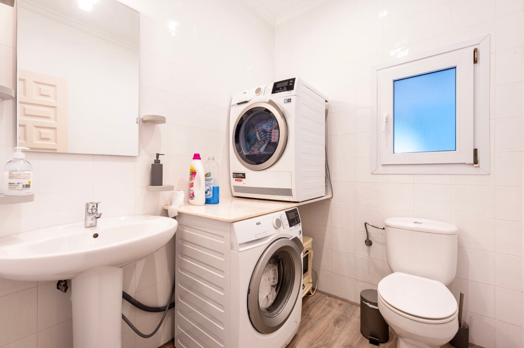 Cuarto de lavado y aseo funcional con paredes alicatadas en blanco, lavabo de pedestal e instalación para lavadora y secadora AEG sobre suelo laminado.
