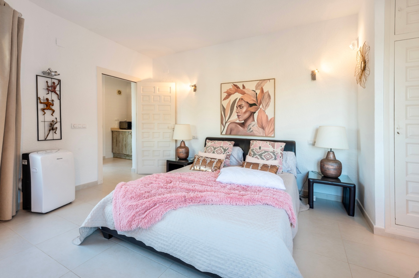 Dormitorio con cama doble, textiles rosados y apliques de pared. Incluye armarios empotrados blancos, aire acondicionado portátil y suelos de gres claro.