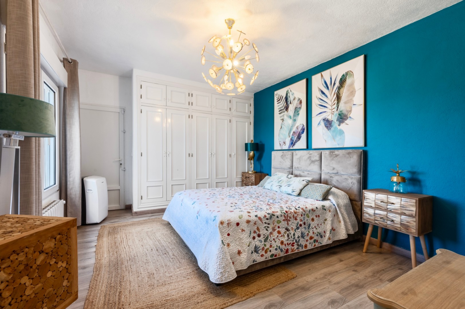 Dormitorio con pared de acento azul turquesa, armarios empotrados blancos y cabecero tapizado. Incluye suelo laminado, alfombra de yute y una moderna lámpara de techo dorada.