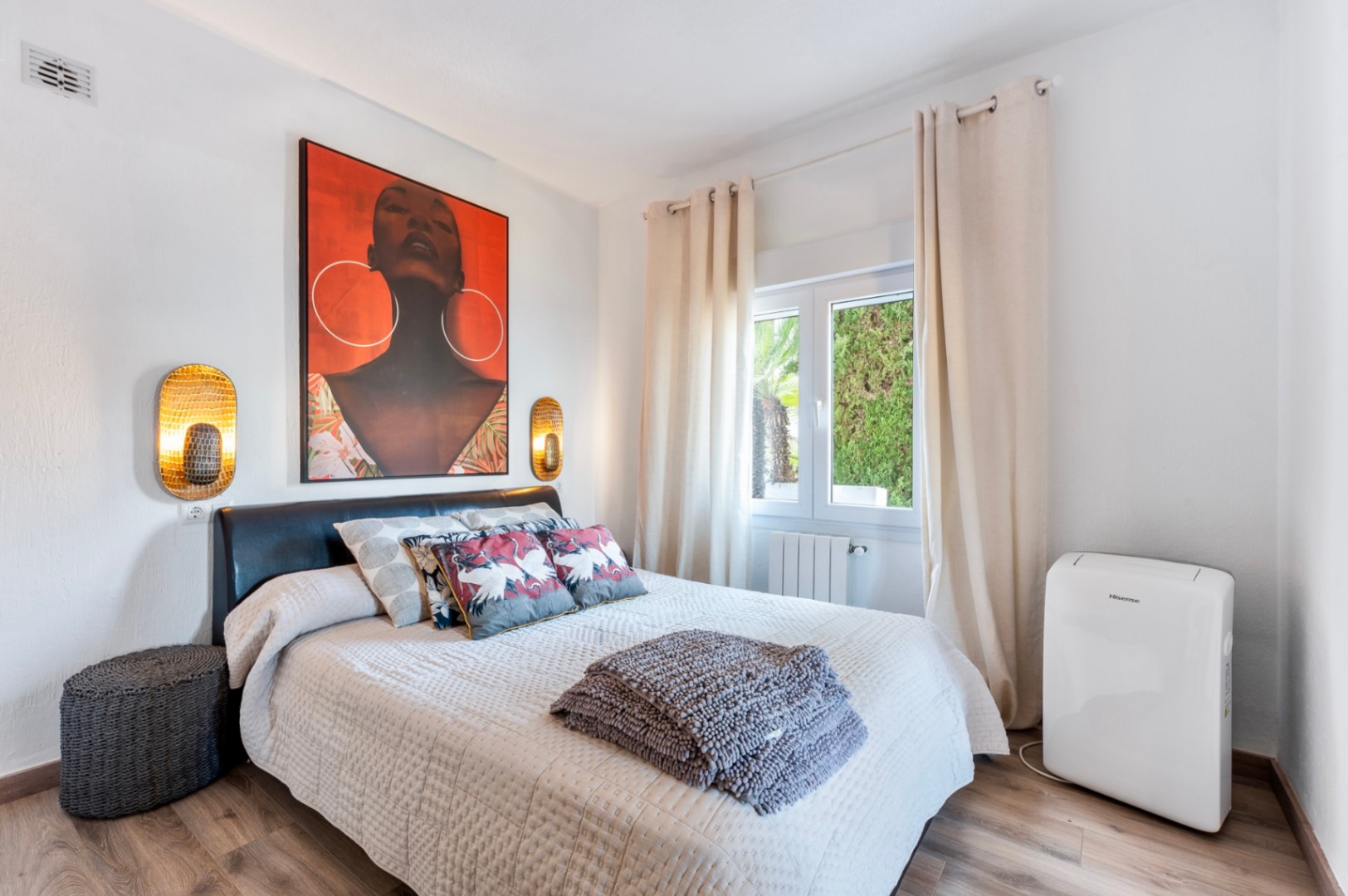 Dormitorio luminoso con suelos de madera, cama doble con cabecero oscuro, apliques dorados y un gran cuadro decorativo. Incluye aire acondicionado portátil y vistas al jardín.