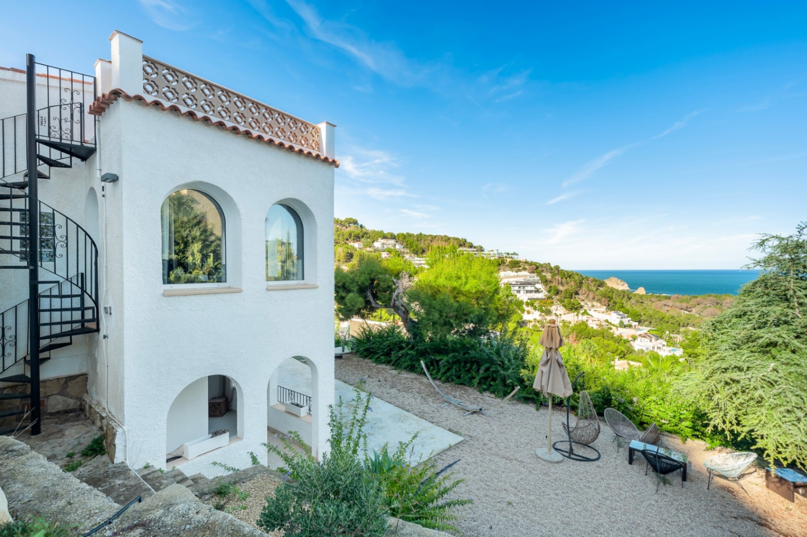 Fachada blanca con arcos tradicionales, escalera de caracol metálica y zona de descanso sobre grava con vistas panorámicas al mar Mediterráneo en Portitxol.