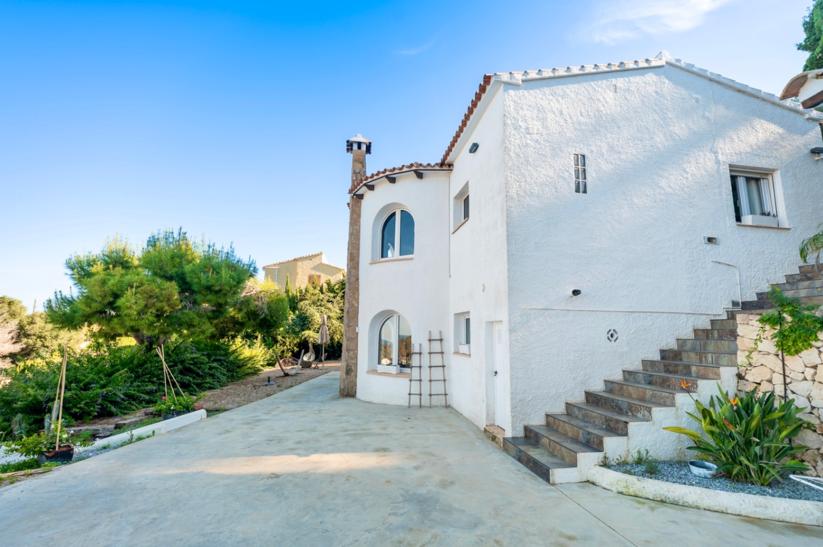 Fachada exterior de la villa con paredes blancas texturizadas, ventanas en arco y una escalera de piedra que conduce a la planta superior, rodeada de vegetación mediterránea.