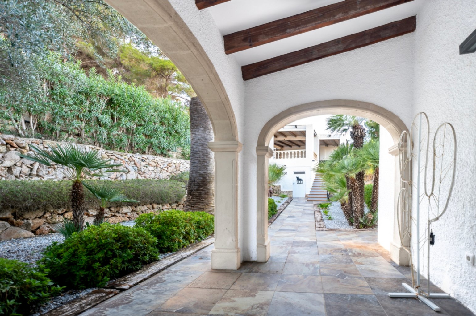 Porche cubierto con arcos de piedra tradicional, vigas de madera vista y suelos cerámicos, con vistas a un jardín mediterráneo con palmeras y muros de piedra natural.