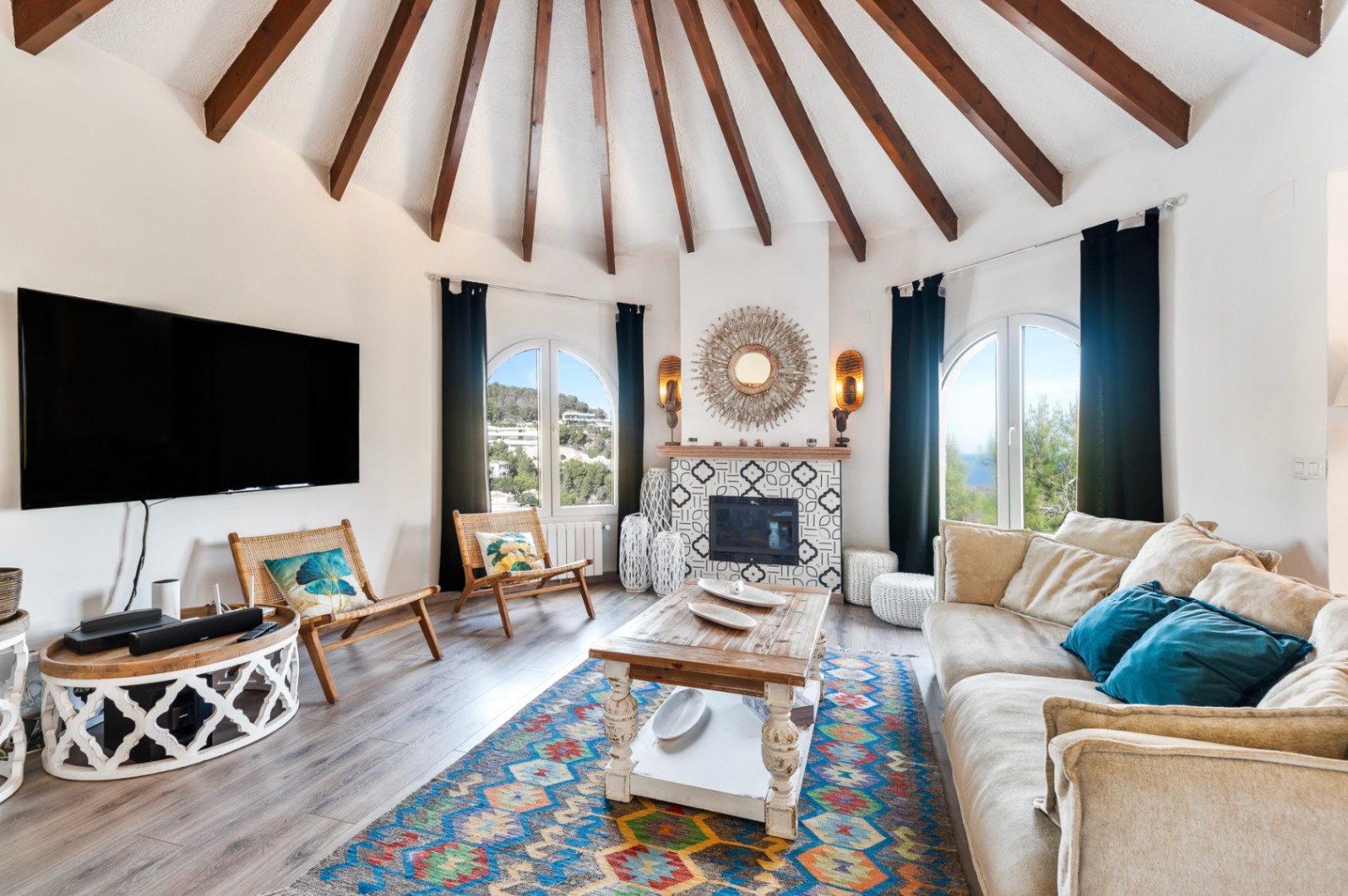 Salón circular con techo de vigas de madera radiantes, chimenea decorada con azulejos geométricos y ventanales en arco con vistas al entorno natural de Jávea.