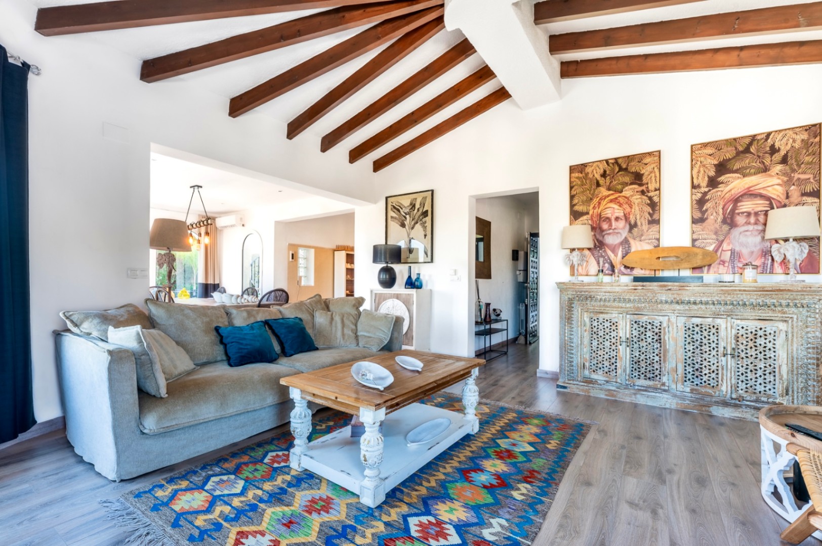 Salón luminoso con techos de vigas de madera vista, mobiliario de estilo rústico y un aparador tallado artesanalmente bajo retratos decorativos.