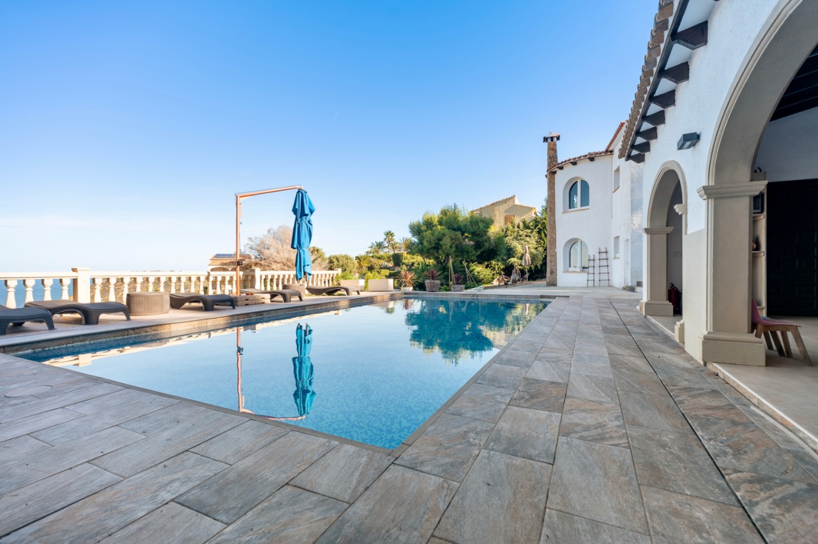 Terraza exterior con piscina rectangular, suelo de piedra natural y vistas al mar. La fachada blanca destaca por sus arcos de estilo tradicional.