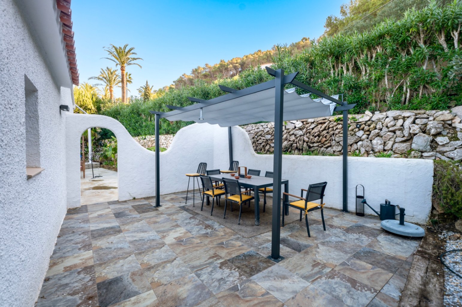 Terraza exterior con suelo de piedra natural y zona de comedor bajo una pérgola moderna. El espacio cuenta con muros de mampostería y vegetación mediterránea.