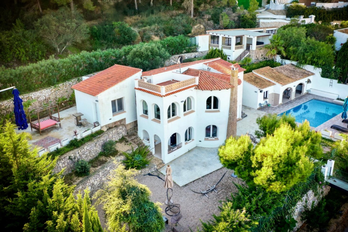 Vista aérea de la villa mediterránea con fachada blanca, tejados de teja roja y ventanas de arco. Destacan la piscina privada, la cocina exterior y los muros de piedra natural.
