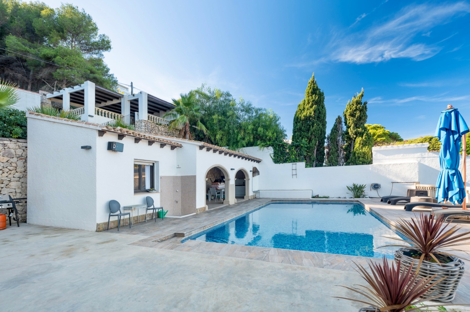 Zona de piscina con terraza de piedra, ducha exterior y porche con arcos. La fachada blanca destaca bajo el cielo despejado, rodeada de vegetación mediterránea y pinos.