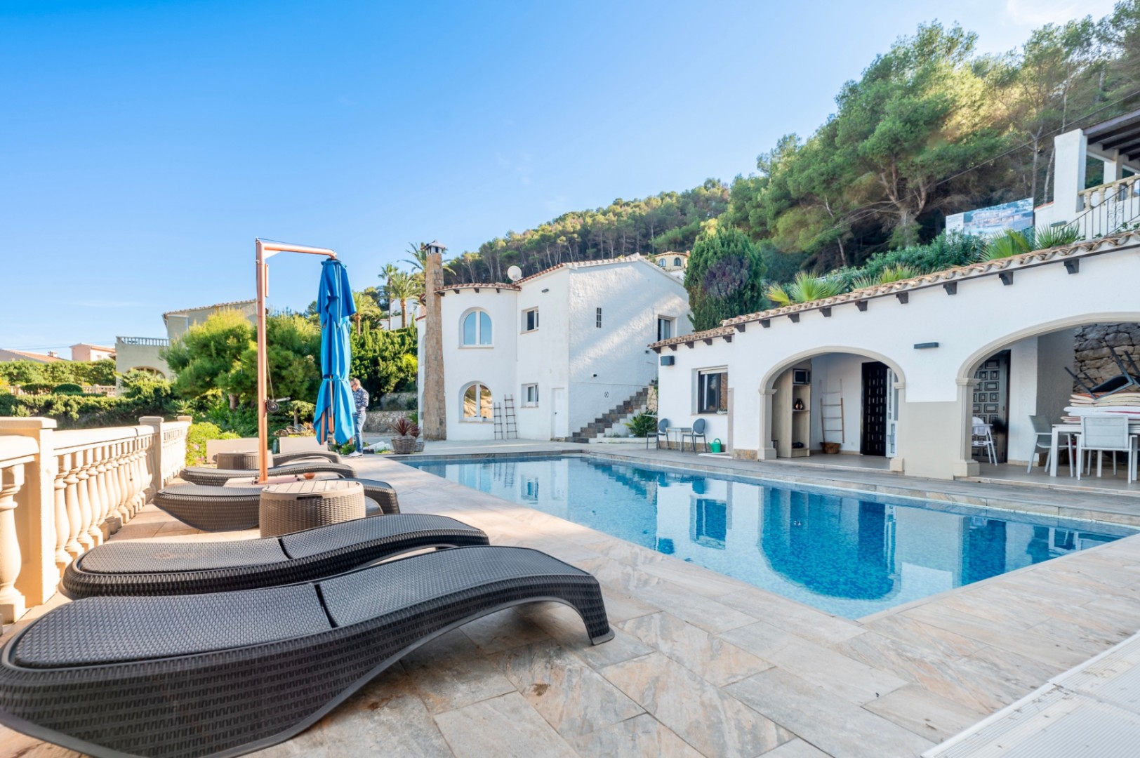 Zona de piscina con tumbonas de ratán, terraza de piedra natural y porches de estilo mediterráneo con arcos frente a una ladera arbolada.