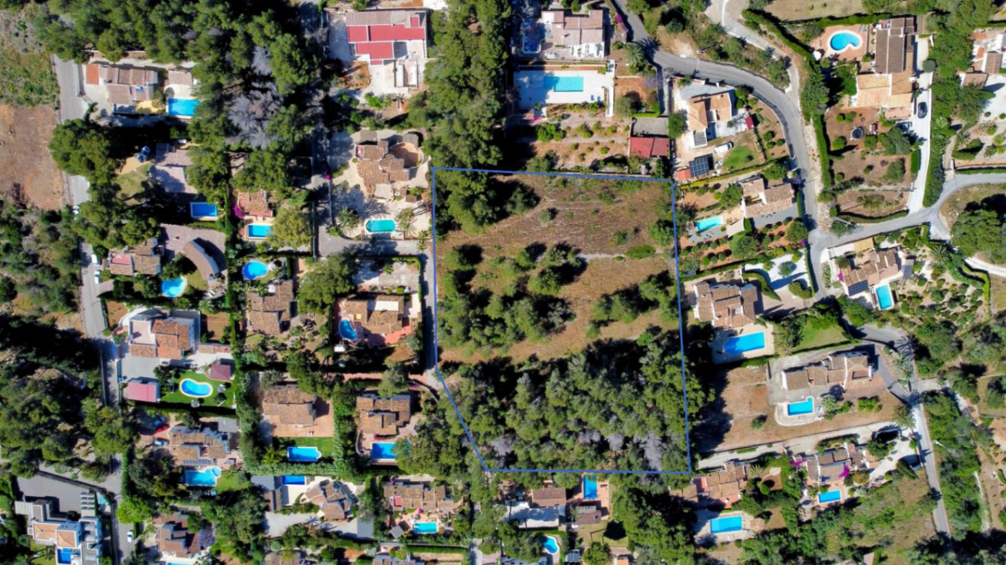 Vista aérea de la parcela llana delimitada en azul, rodeada de villas residenciales con piscinas y abundante vegetación mediterránea.