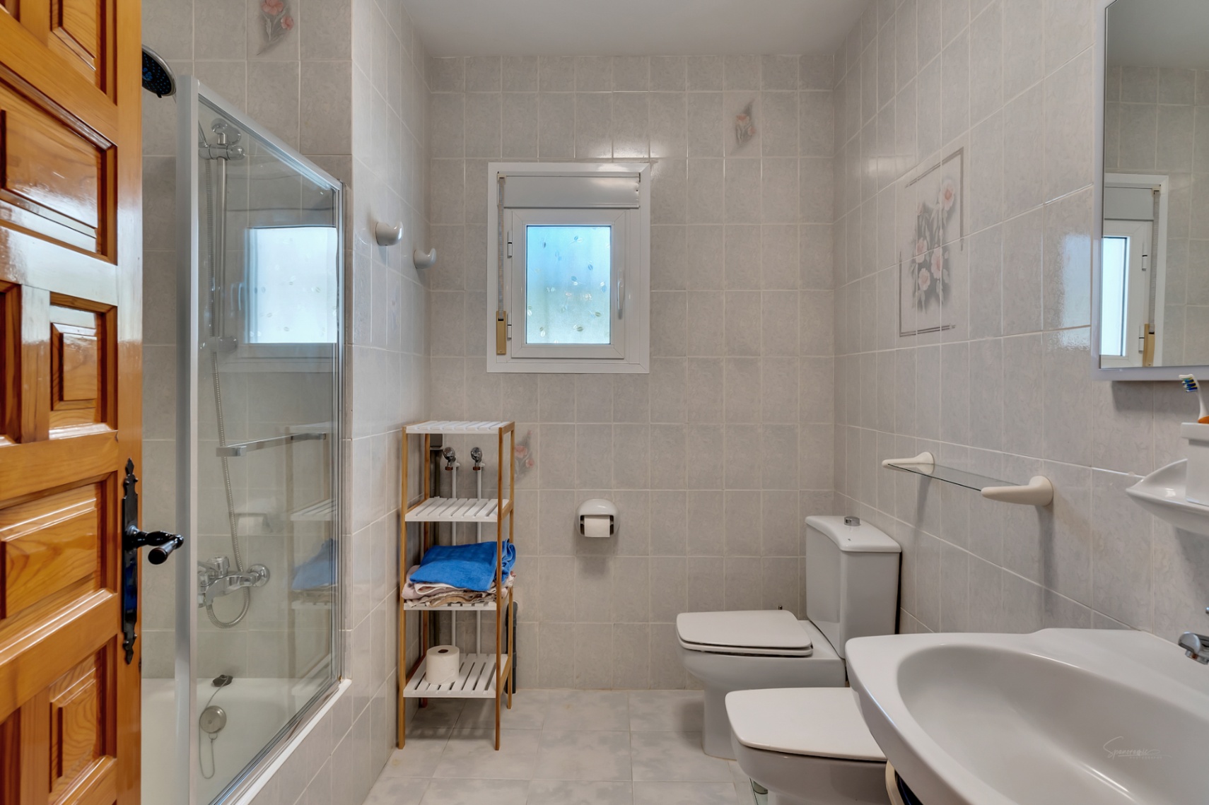 Baño funcional con bañera y mampara de cristal, sanitarios blancos, estantería de madera y paredes alicatadas con detalles florales decorativos.