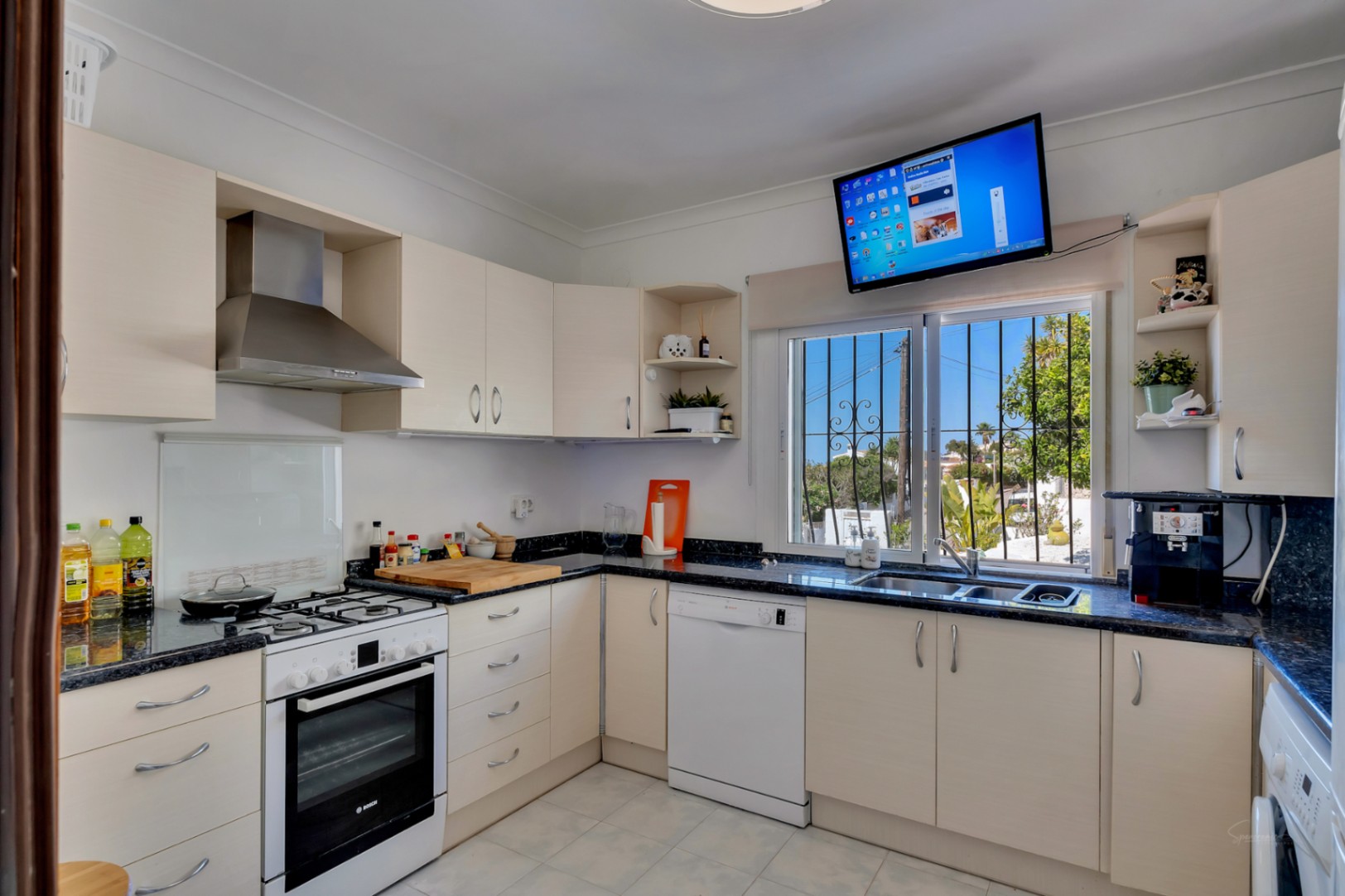 Cocina funcional en forma de L con encimeras de granito oscuro, armarios crema, electrodomésticos blancos y ventana con vistas al exterior y televisor superior.