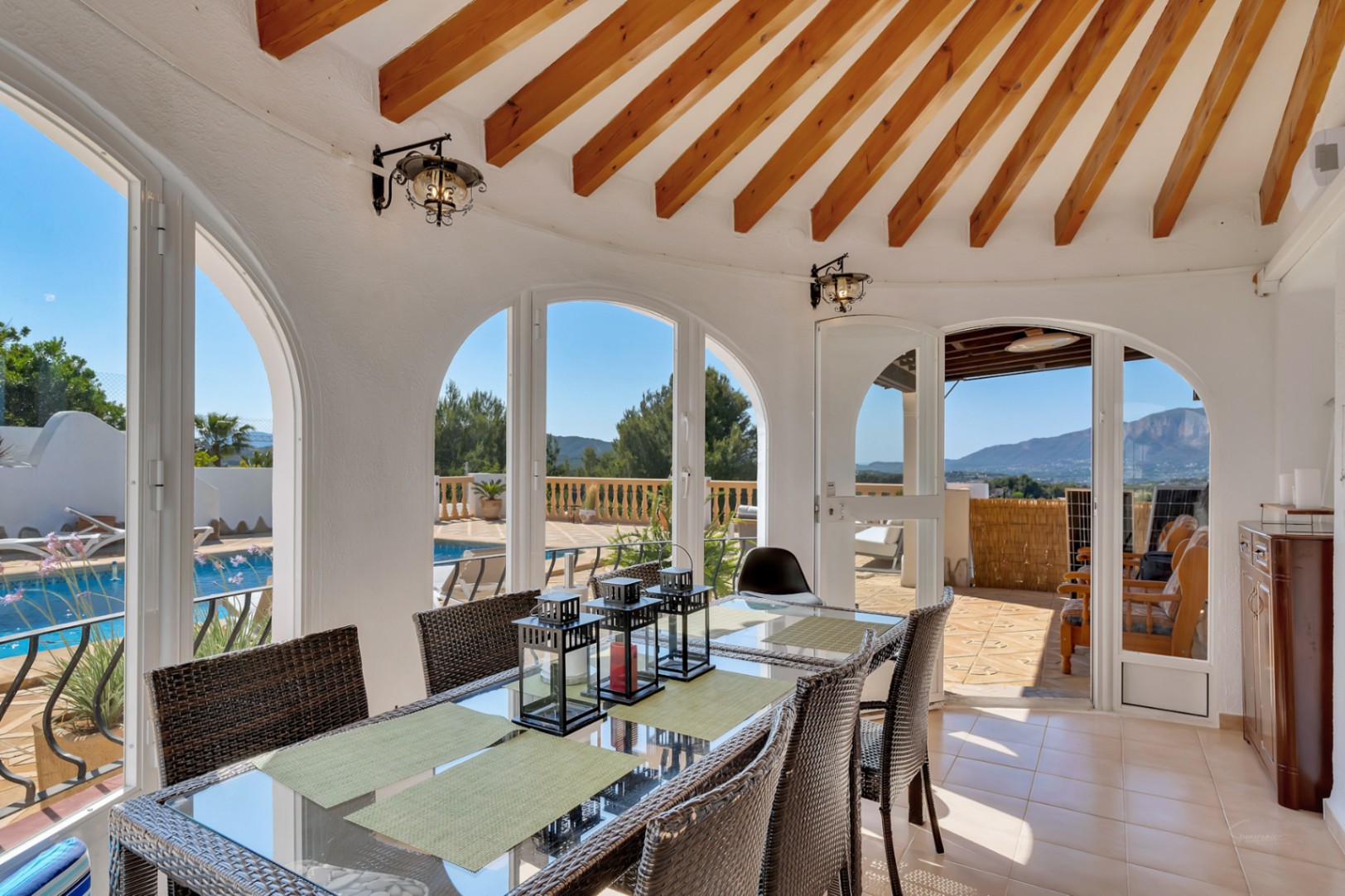 Comedor en naya acristalada con vigas de madera radiales, mesa de cristal y vistas directas a la piscina y al Montgó a través de sus arcos.