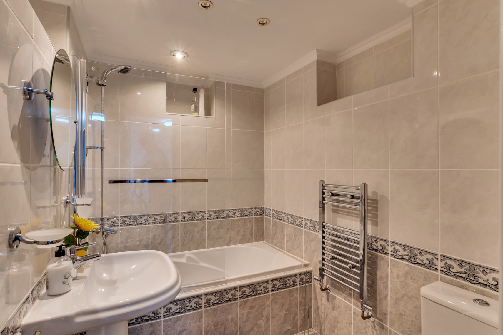 Cuarto de baño alicatado con bañera integrada, mampara de cristal, lavabo de pedestal, radiador toallero cromado e iluminación mediante focos empotrados.