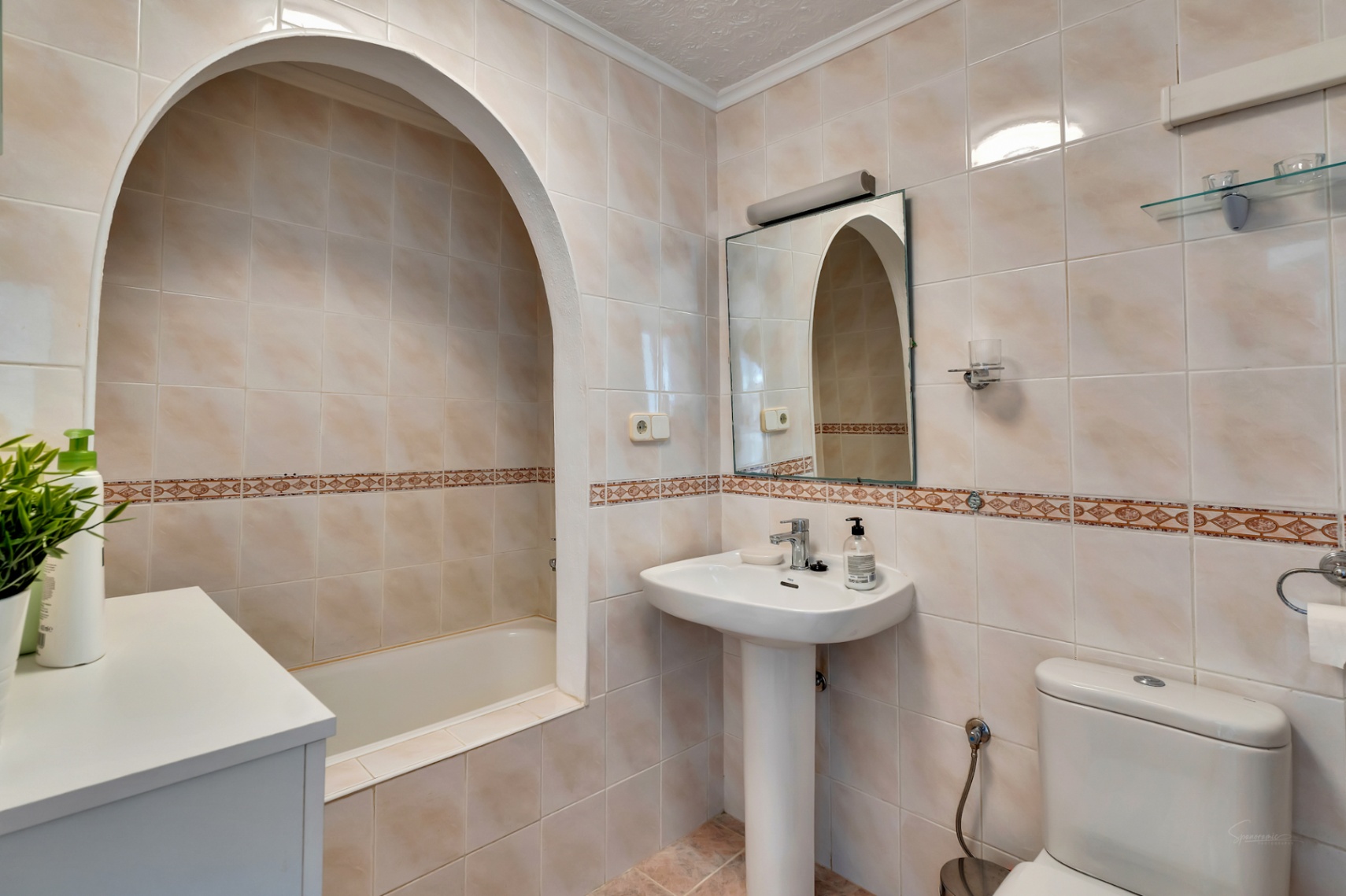 Cuarto de baño con bañera encastrada bajo un arco decorativo, lavabo de pedestal y paredes revestidas en azulejos cerámicos con cenefa ornamental.