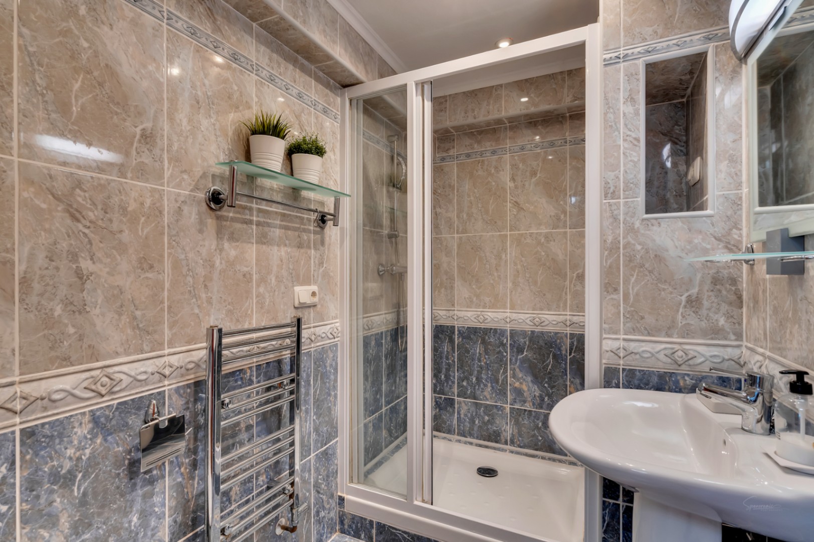 Cuarto de baño con ducha de mampara blanca, azulejos de efecto mármol en tonos beige y azul, radiador toallero cromado y lavabo de pedestal.