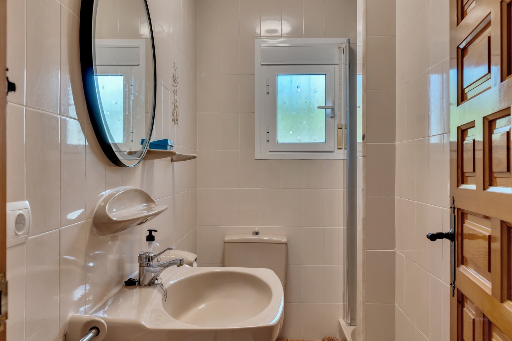 Cuarto de baño funcional con azulejos en tonos crema, espejo circular de marco negro, lavabo cerámico y ventana oscilobatiente para ventilación natural.