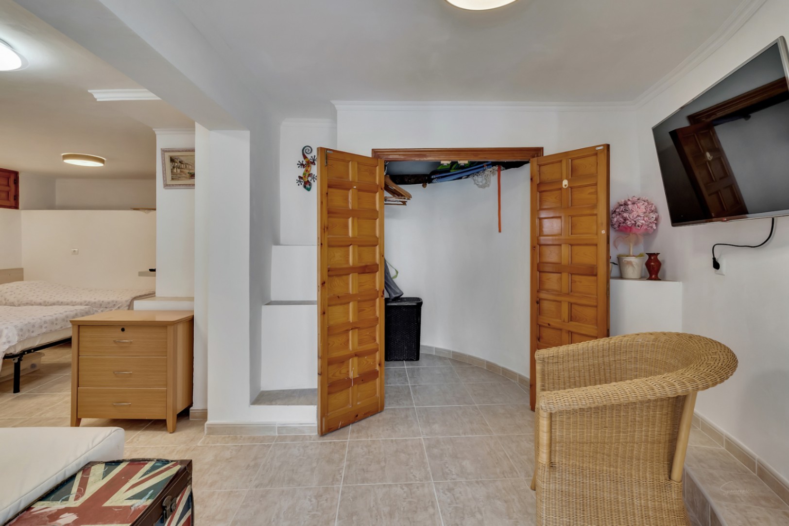 Dormitorio con armario empotrado de madera, suelos cerámicos y zona de estar con sillón de mimbre y televisión de pantalla plana.