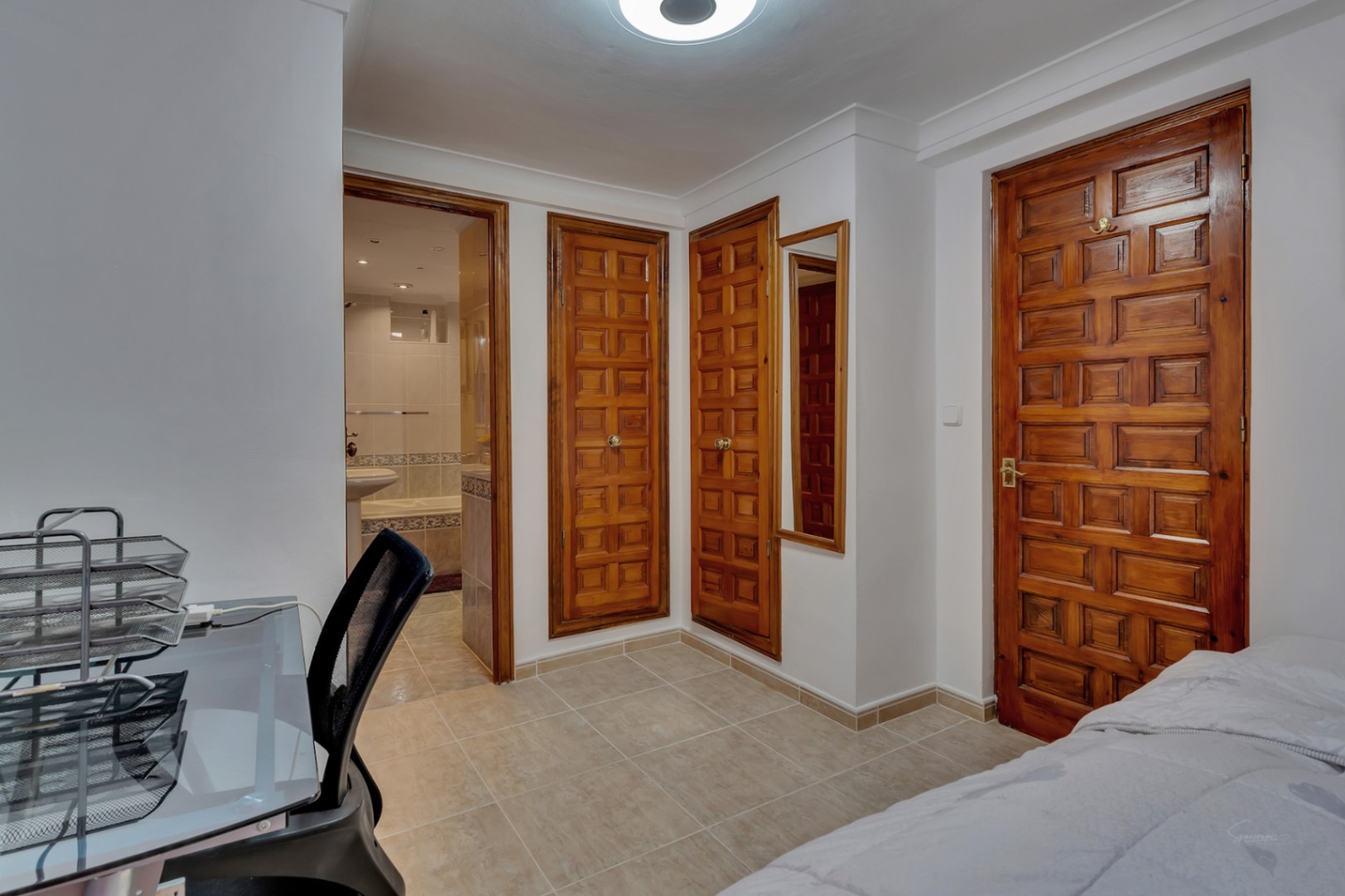Dormitorio con armarios empotrados de madera tallada, suelos cerámicos y acceso directo a un baño en suite con bañera y acabados en tonos neutros.