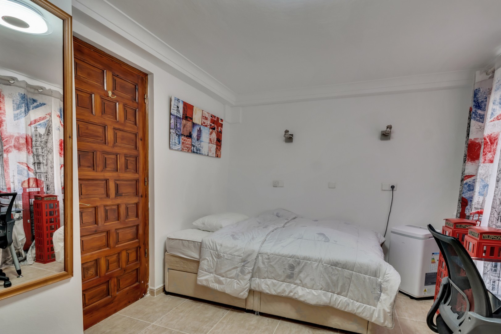 Dormitorio con cama doble, suelos cerámicos y puerta de madera tallada. La estancia cuenta con iluminación de pared y detalles decorativos de temática urbana.