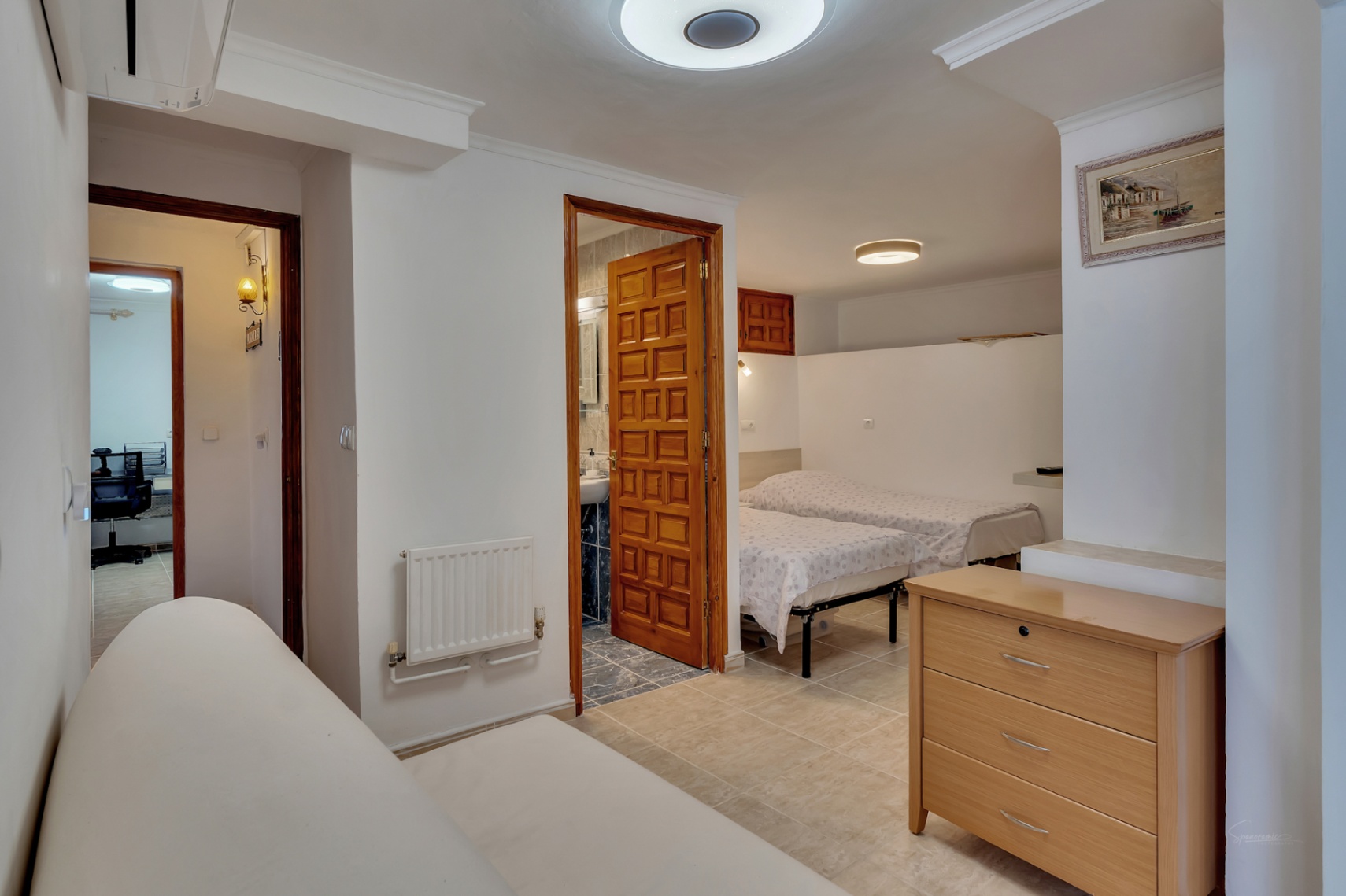 Dormitorio con dos camas individuales, baño en suite con puerta de madera artesanal, radiador y suelos cerámicos. Al fondo se aprecia un acceso a zona de despacho.