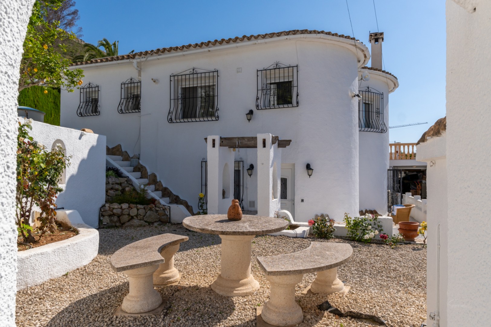 Fachada blanca de estilo mediterráneo con mesa y bancos de piedra en un patio de grava, rodeado de vegetación y detalles de forja en las ventanas.