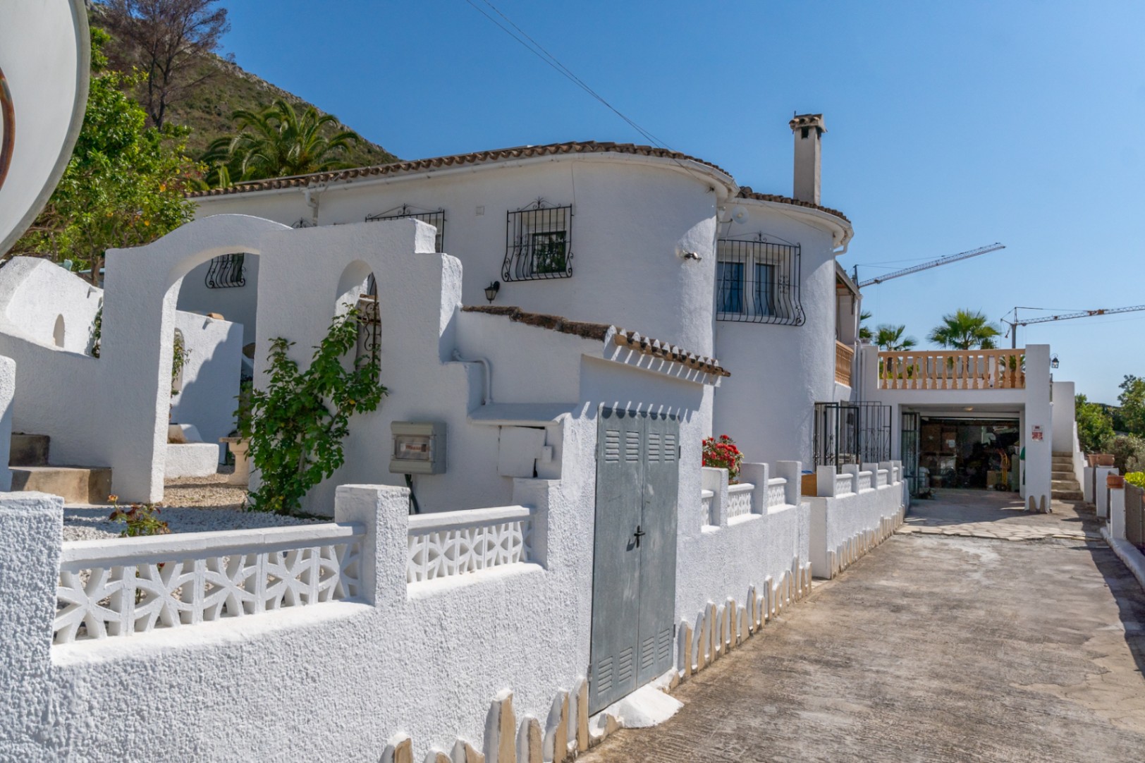 Fachada exterior de estilo mediterráneo con muros blancos texturizados, detalles de arcos, carpintería con rejas de hierro y acceso lateral hacia la zona de garaje.