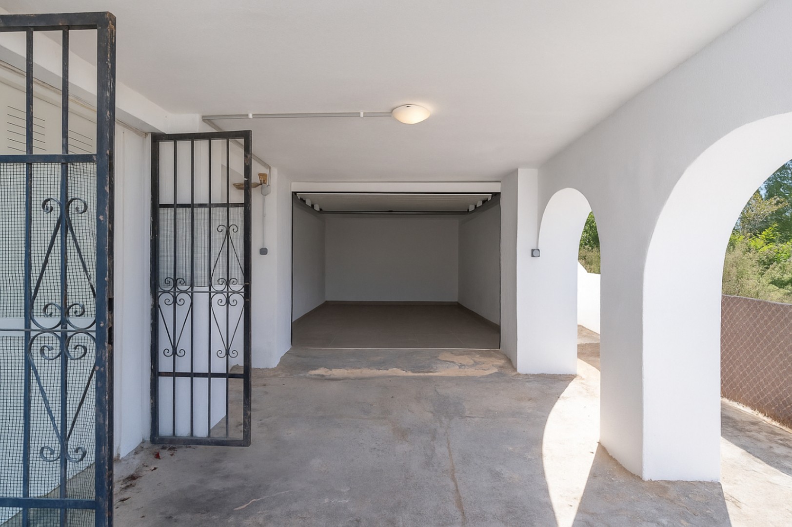 Garaje cerrado y zona de aparcamiento cubierta con arcos mediterráneos, paredes blancas y puerta de seguridad de forja negra.