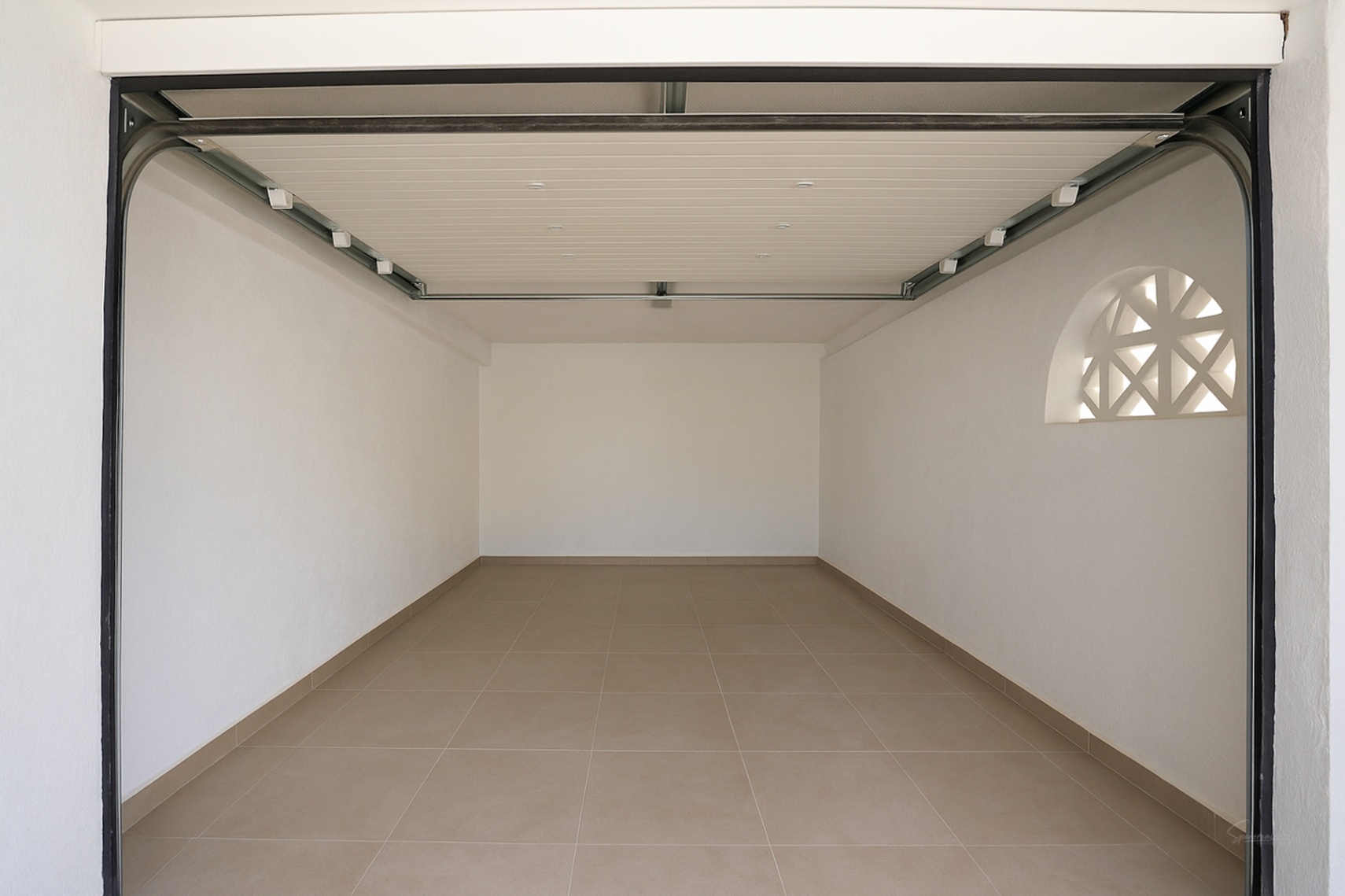 Garaje privado con suelo cerámico, paredes blancas y puerta seccional automática. Incluye una ventana decorativa de arco con celosía para luz natural.