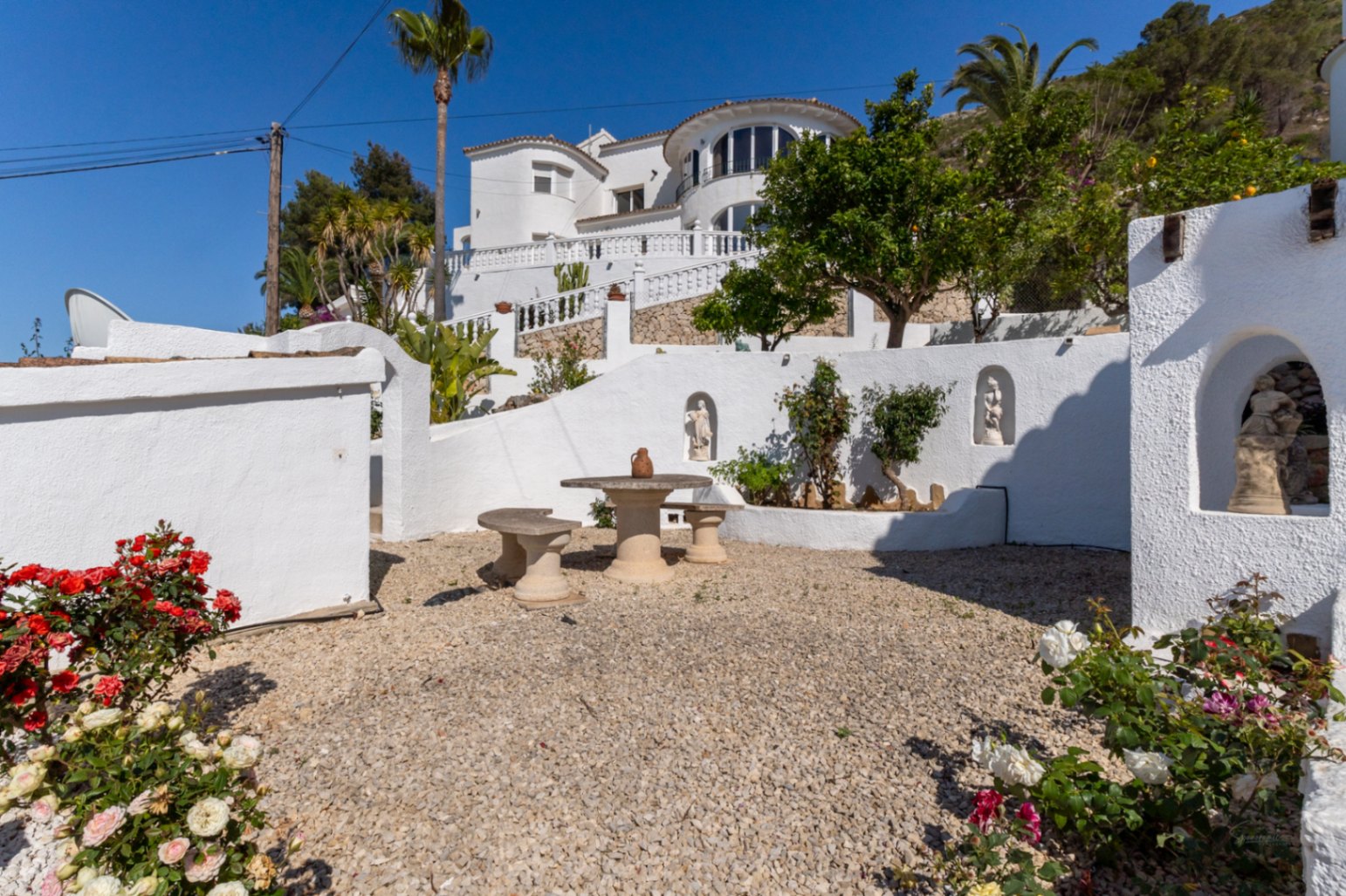 Jardín aterrazado con suelo de grava, mesa de piedra y nichos decorativos en muros blancos, con la fachada principal de la villa al fondo.