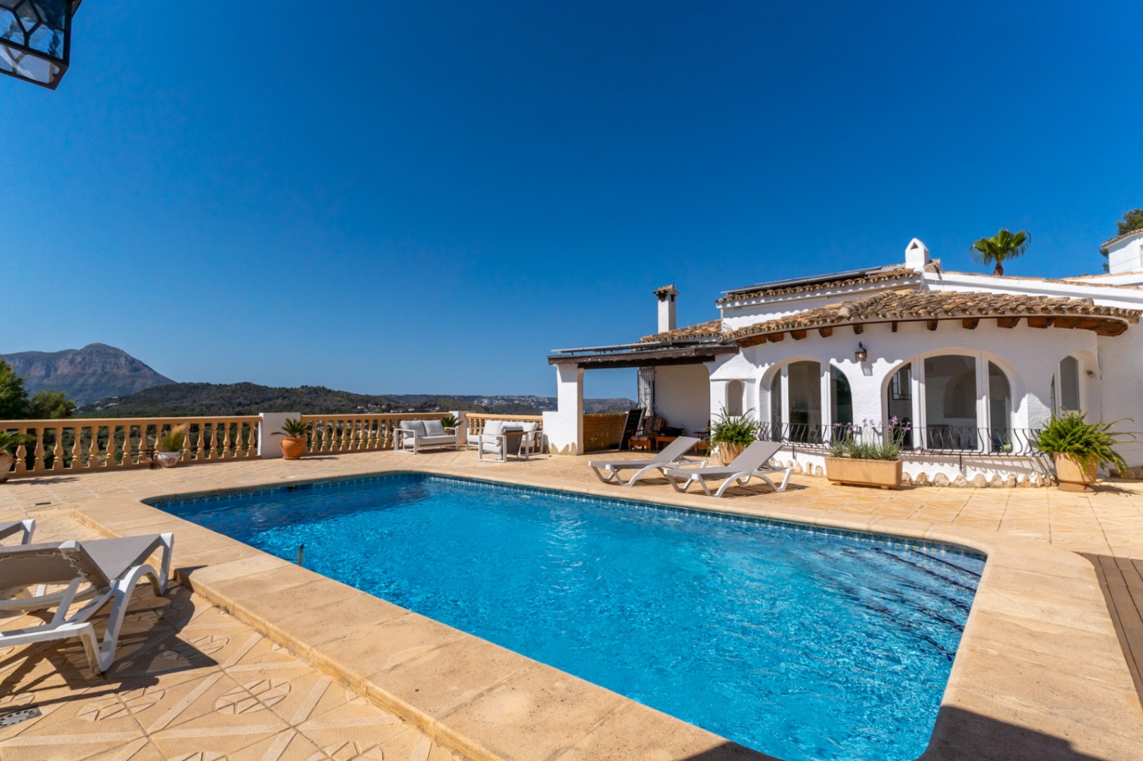 Piscina rectangular rodeada de una amplia terraza de gres con zona de solárium, balaustrada clásica y vistas despejadas al Montgó bajo un cielo despejado.