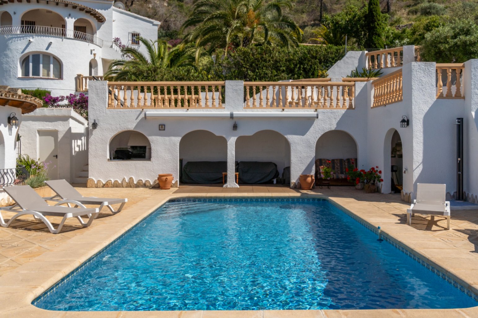 Piscina rectangular rodeada de una terraza de piedra con tumbonas, porches de arcos tradicionales y balaustradas blancas bajo la vegetación mediterránea.