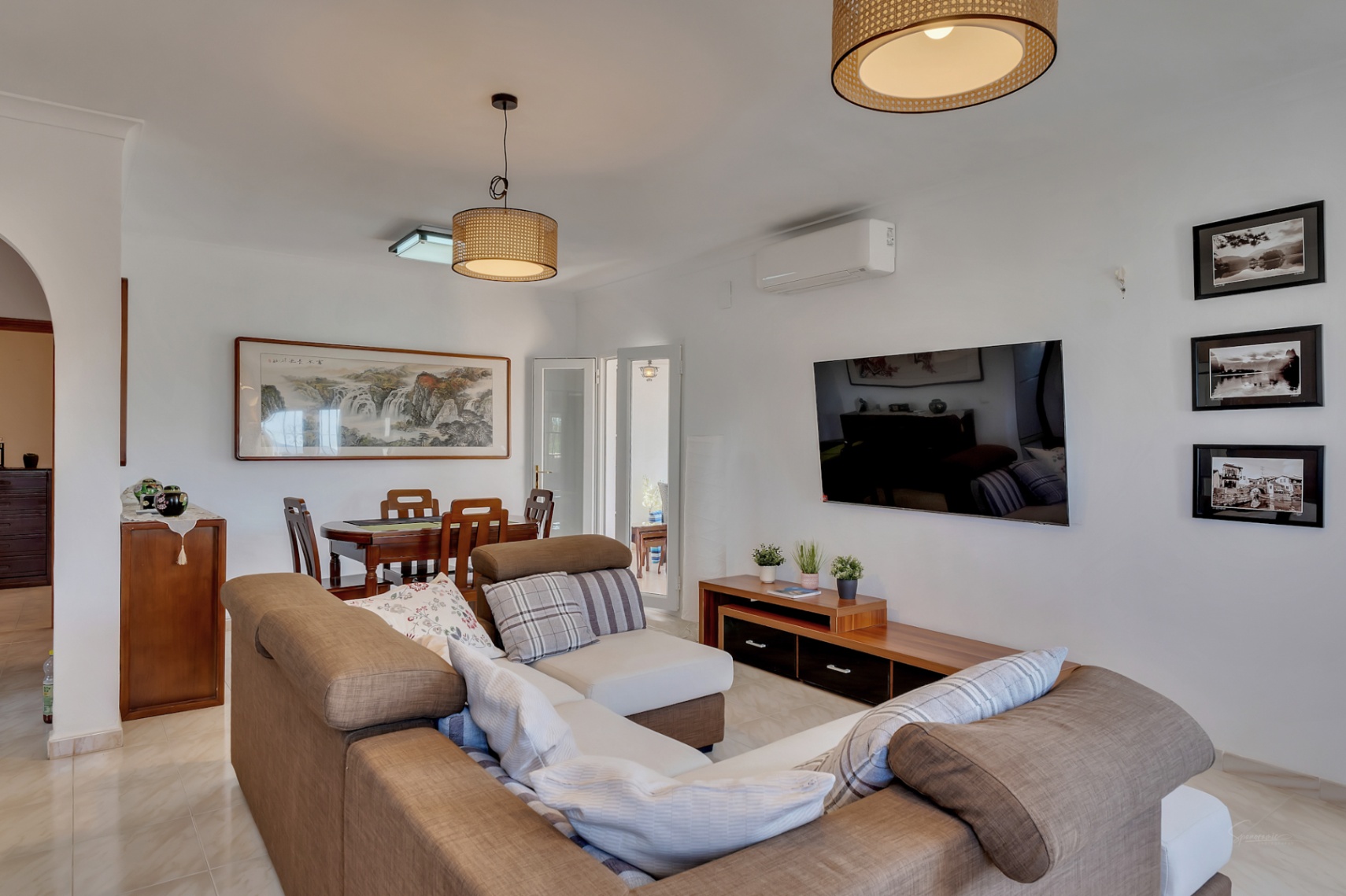 Salón comedor integrado con sofá rinconero en tonos tierra, mesa de comedor de madera y lámparas de fibras naturales. Incluye aire acondicionado y suelos cerámicos claros.