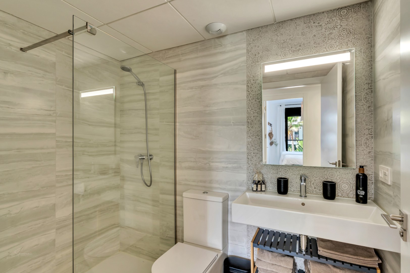 Baño de diseño con ducha de obra, mampara de cristal y azulejos efecto mármol. Incluye un lavabo suspendido sobre una pared de mosaico gris y un espejo con iluminación integrada.