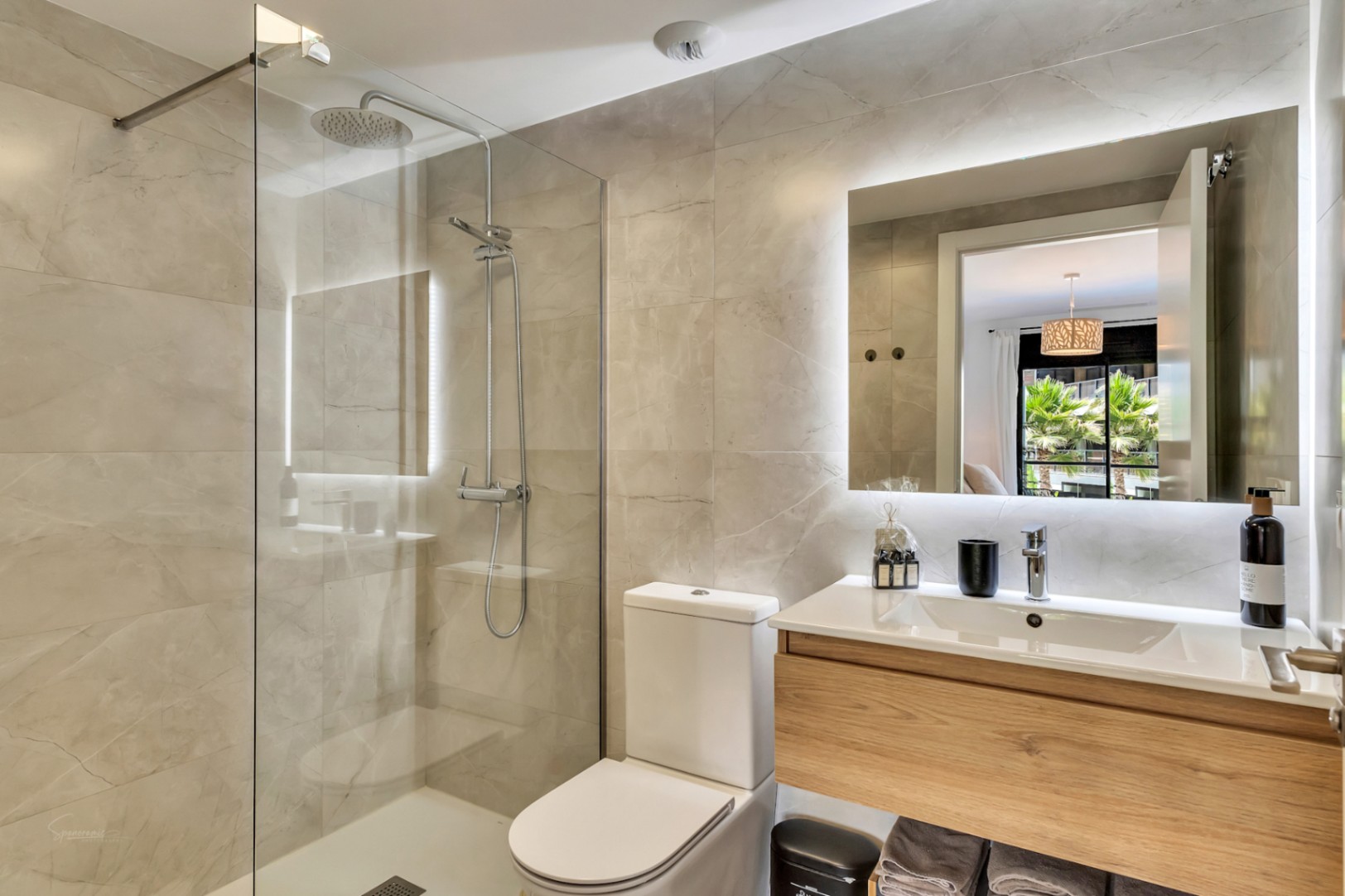 Baño moderno con ducha de efecto lluvia, mampara de cristal y paredes de gres porcelánico. Destaca el mueble suspendido de madera y el espejo con retroiluminación LED integrada.