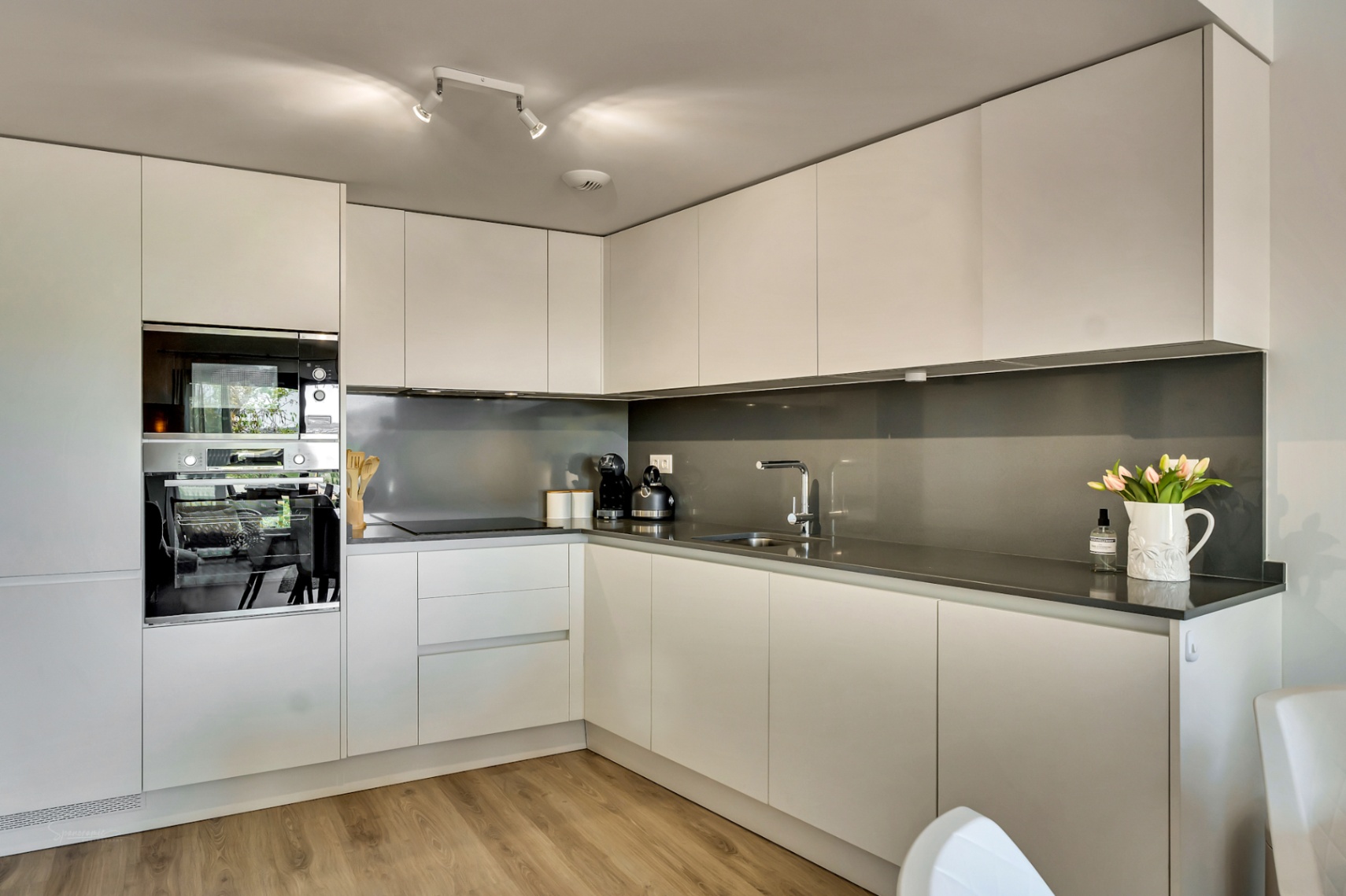 Cocina moderna en L con armarios blancos minimalistas, encimera de cuarzo gris oscuro y electrodomésticos integrados de alta gama sobre suelos de madera clara.