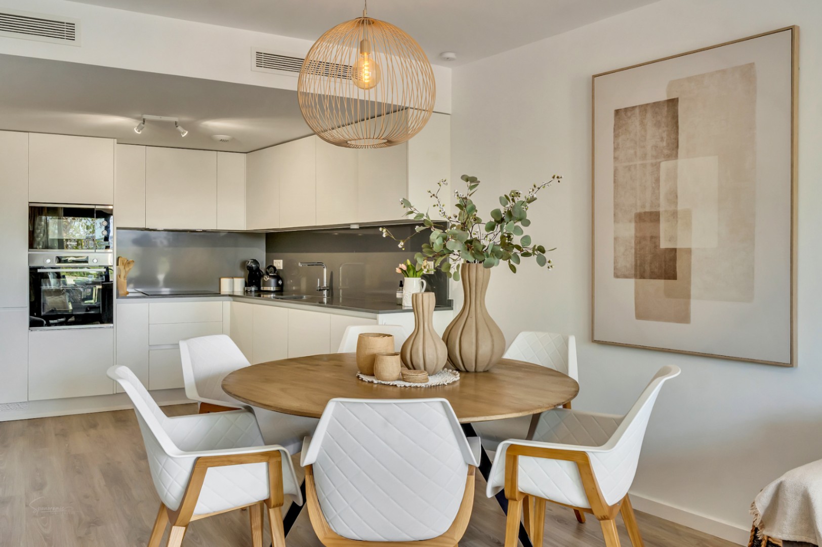 Comedor de concepto abierto con mesa circular de madera, sillas de diseño acolchadas y una lámpara de mimbre, integrado con una cocina blanca moderna y funcional.