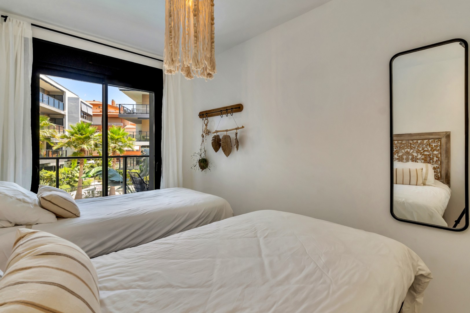 Dormitorio con dos camas individuales, lámpara de flecos y espejo de pared. El ventanal ofrece vistas directas a la urbanización y zonas verdes.