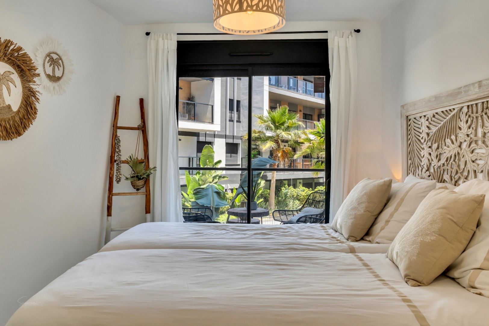 Dormitorio luminoso con cabecero de madera tallada, decoración de estilo mediterráneo y acceso directo a una terraza con vistas a las zonas comunes ajardinadas.