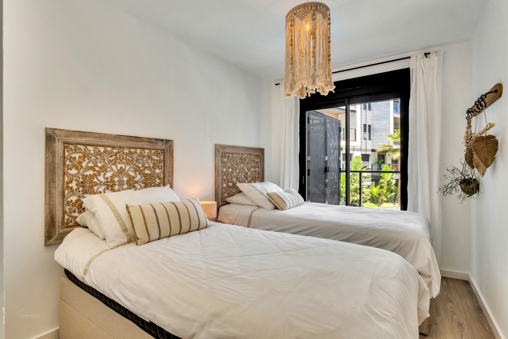 Dormitorio luminoso con dos camas individuales, cabeceros de madera tallada y lámpara de macramé. El ventanal ofrece acceso directo a la terraza con vistas al jardín.