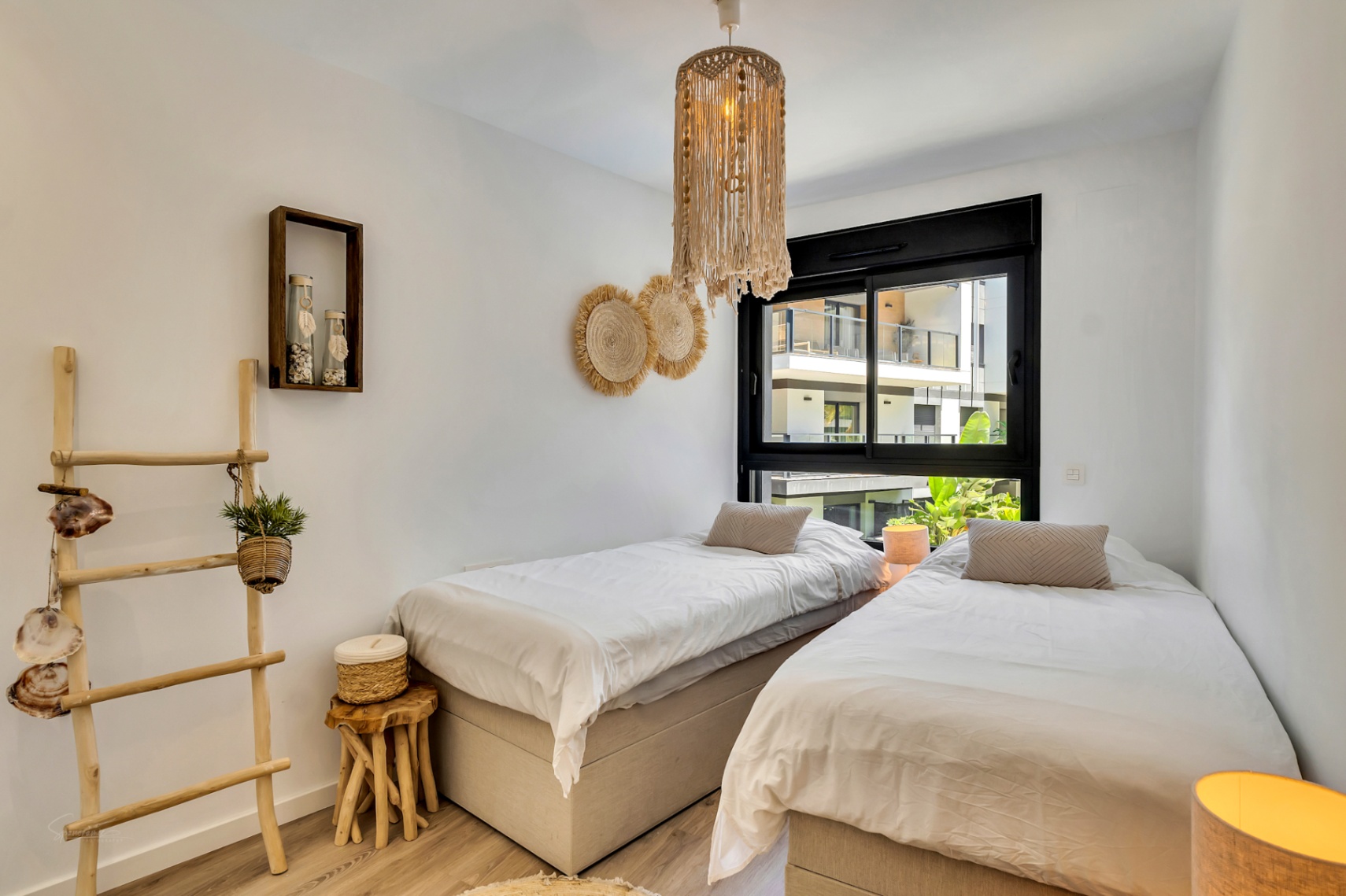Dormitorio luminoso con dos camas individuales, decoración de estilo mediterráneo con elementos de madera natural, lámpara de macramé y ventana con carpintería negra.