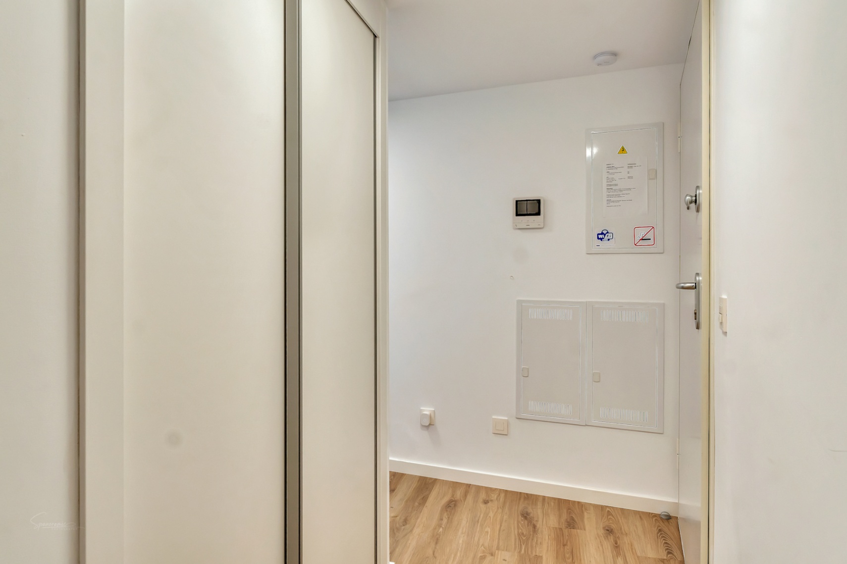 Pasillo de entrada con suelos de madera clara, armario empotrado blanco de gran capacidad y paneles de control integrados en la pared para mayor funcionalidad.