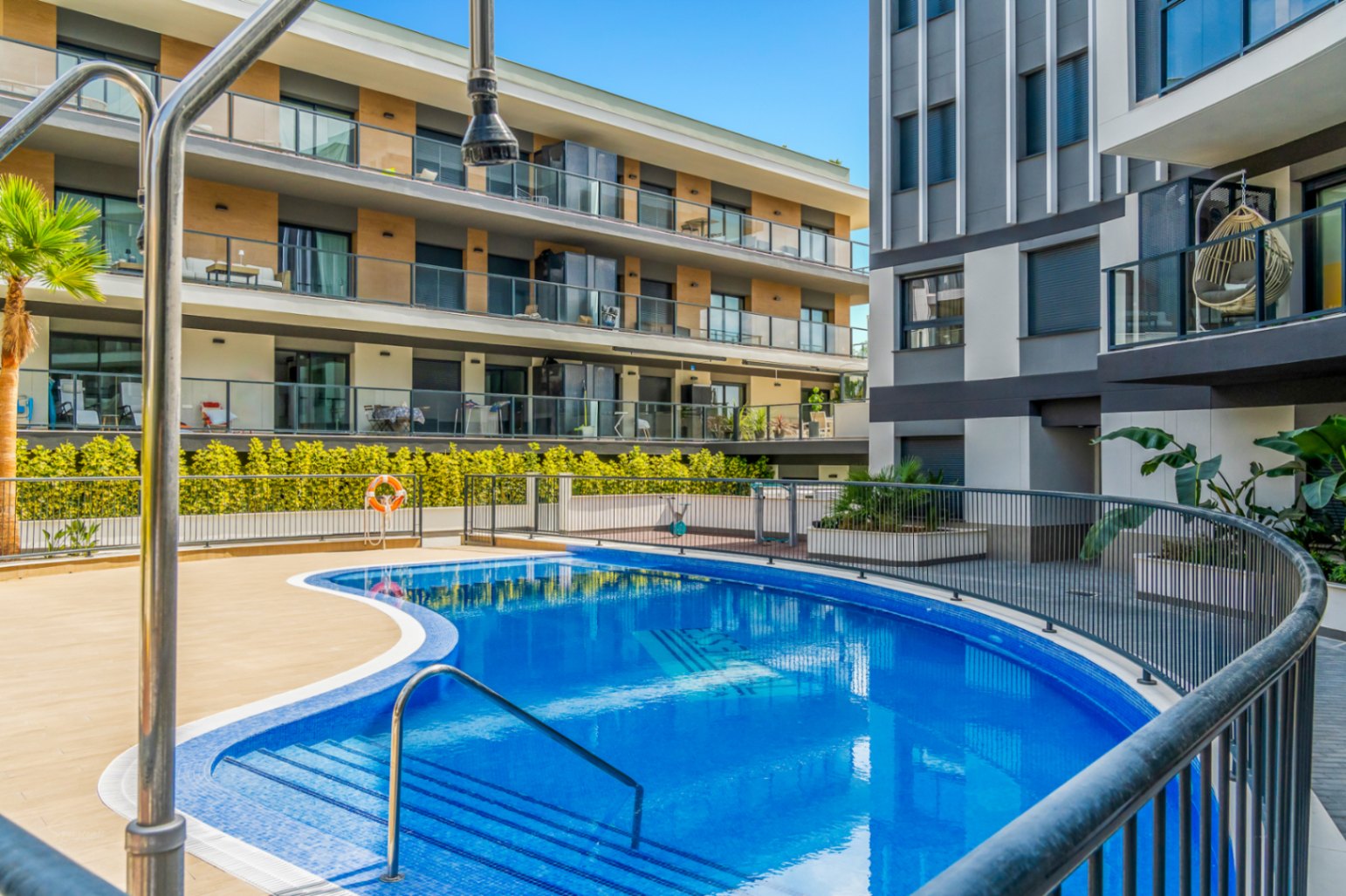 Piscina comunitaria de diseño curvo rodeada de una amplia terraza y edificios residenciales modernos con balcones de cristal y acabados en madera.
