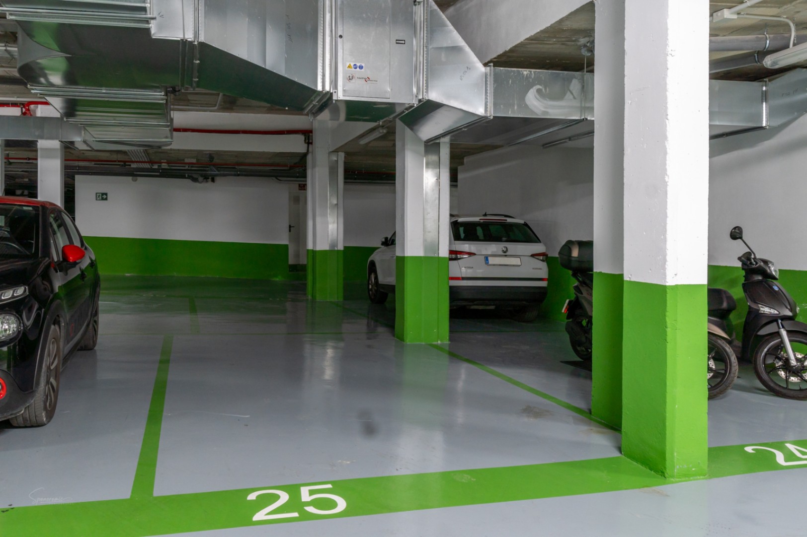 Plaza de garaje subterránea numerada con suelo de resina gris y señalización en verde. Incluye columnas de hormigón y conductos de ventilación vistos.