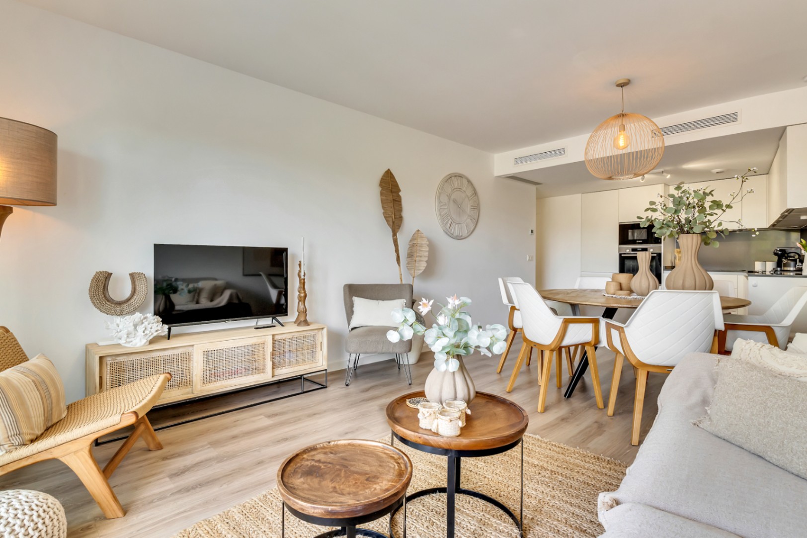 Salón-comedor de concepto abierto con suelos de madera, mobiliario de estilo natural y cocina integrada al fondo con electrodomésticos empotrados.