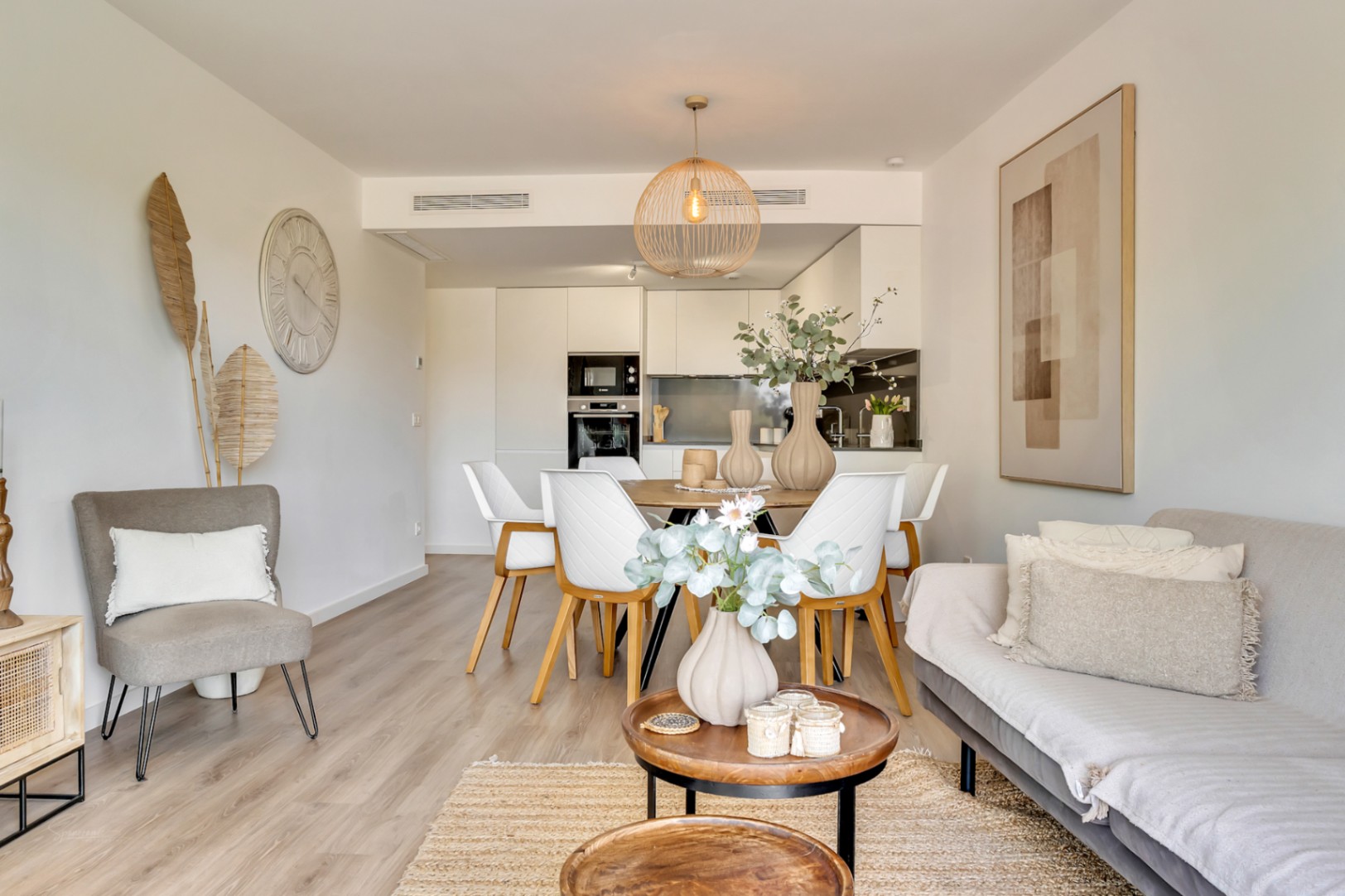 Salón-comedor diáfano con suelos de madera clara, mobiliario de tonos neutros y cocina integrada. Destaca la iluminación natural y el diseño contemporáneo con detalles en fibras naturales.